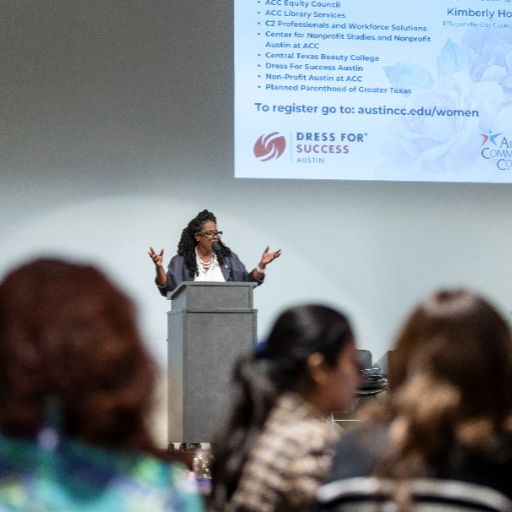 A Black woman wearing a white top and dark blazer speaks to the crowd at the ACC Women’s Health, Motivation, and Empowerment Conference about the nonprofit Dress for Success