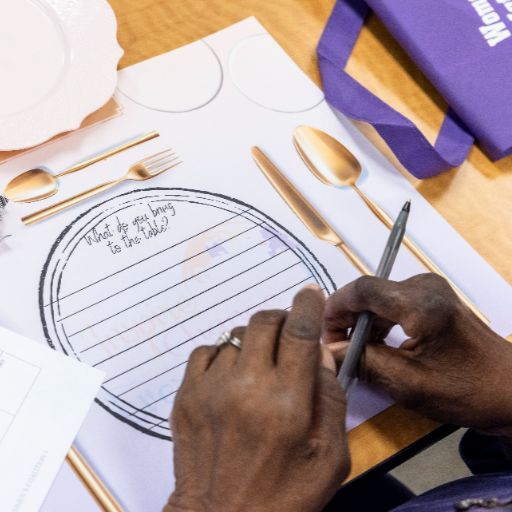 The hands of a Black person rests on a table setting, ready to write in answer to the prompt, what do you bring to the table? At the ACC Women’s Health, Motivation, and Empowerment Conference