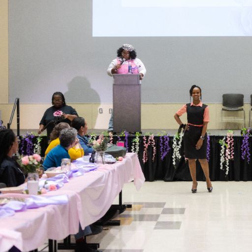 A speaker wearing a cute cap and a pink top stands at a dais, introducing fashion show models wearing business casual outfits from Dress for Success while attendees at a pink draped table watch at the ACC Women’s Health, Motivation, and Empowerment Conference