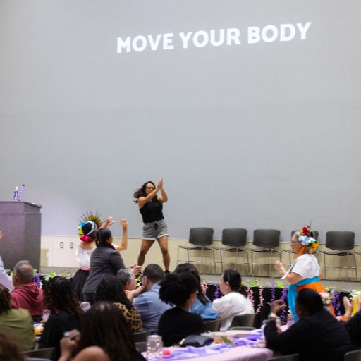 A woman wearing a black t shirt and shorts demonstrates a dance move to attendees at the ACC Women’s Health, Motivation, and Empowerment Conference