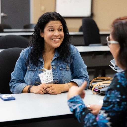 A woman with dark hair wearing a jean jacket smiles at a woman with glasses, brown hair, and a blue patterned shirt at the ACC Women’s Health, Motivation, and Empowerment Conference