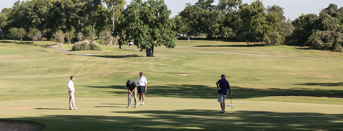 A scenic view of a golf course on a sunny day. In the foreground four golfers are engaged in a game, with one player putting on the green while the others observe. Lush green trees and grass surround the area, creating a peaceful outdoor atmosphere.