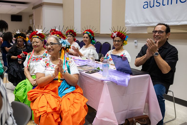 Attendees wearing Mexican and Latin American traditional costumes in bright skirts, white embroidered blouses, and crowns of flowers applaud at the community conference.